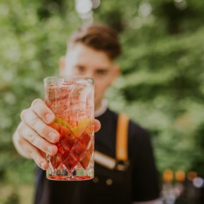 Ein Mann h&auml;lt ein Glas mit rotem Getr&auml;nk und Eisw&uuml;rfeln vor gr&uuml;nem Hintergrund im Hotel Klosterhof.