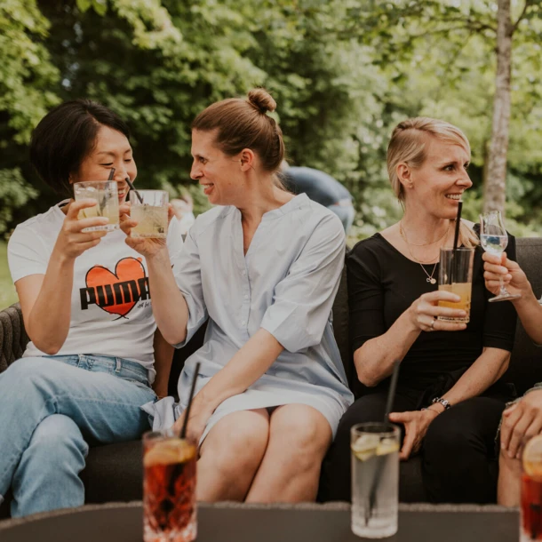 Four women enjoy drinks on a garden sofa, laughing and toasting in a relaxed outdoor setting.